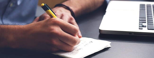 Ruka zapisuje poznámky na papír vedle otevřeného notebooku.