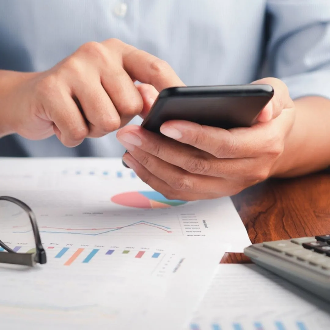 Osoba používá mobilní telefon nad finančními přehledy a kalkulačkou.